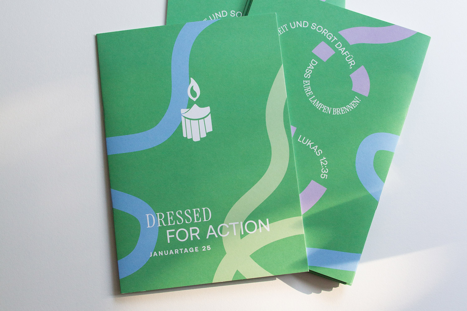 Zwei grüne Broschüren mit abstrakten blauen und hellgrünen Linien, weiße Schrift ‚Dressed for Action Januartage 25‘ und eine Kerzenillustration.