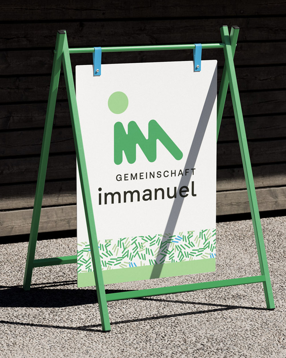Grünes Aufsteller-Schild mit dem Logo und Schriftzug der Gemeinschaft Immanuel vor dunkler Holzfassade.