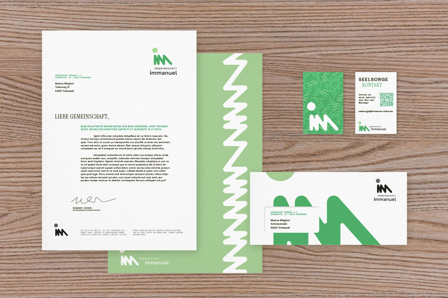 Briefbogen, grüne Mappe, Visitenkarte, Kontaktkarte mit QR-Code und Umschlag mit grünem Design der Gemeinschaft Immanuel auf Holztisch.