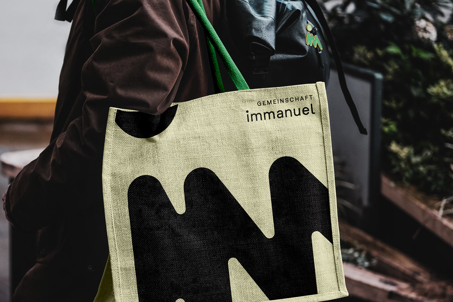 Eine Person trägt eine beige Stofftasche mit grünem Griff, bedruckt mit dem Schriftzug 'Gemeinschaft immanuel' und einem großen schwarzen Muster.