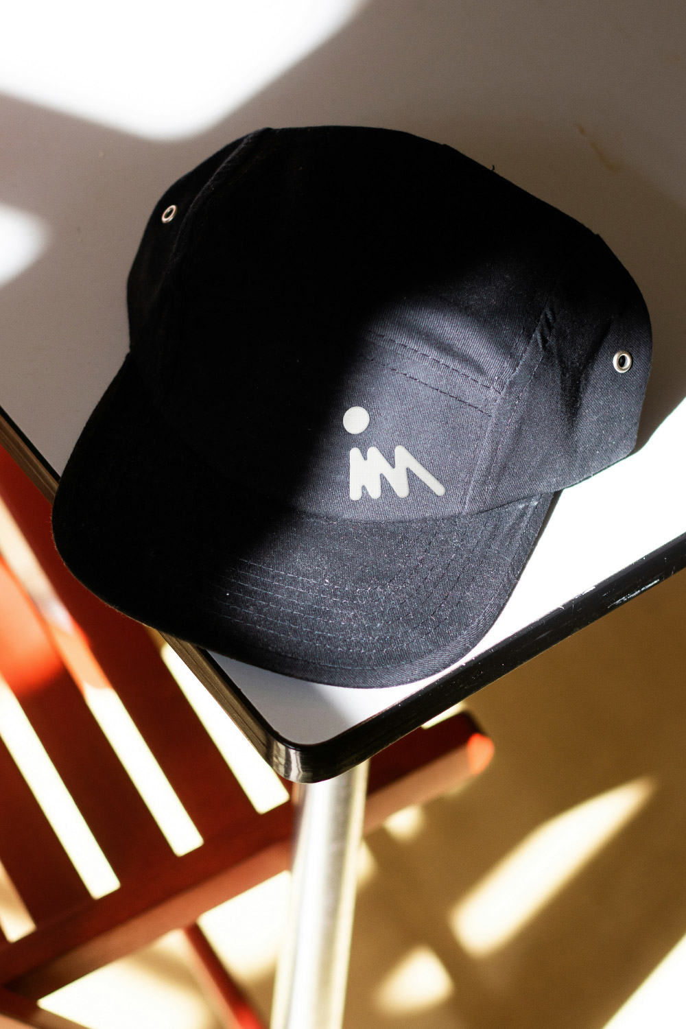 Schwarze Kappe mit einem weißen Logo, liegend auf der Ecke eines Tisches mit Sonnenlicht und Schatten.