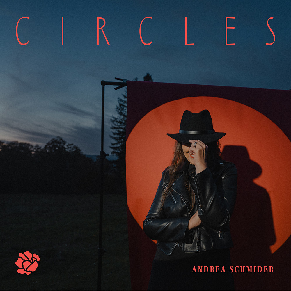 Frau in schwarzem Lederjacke und Hut steht vor orangefarbenem Kreis vor dunklem Abendhimmel.