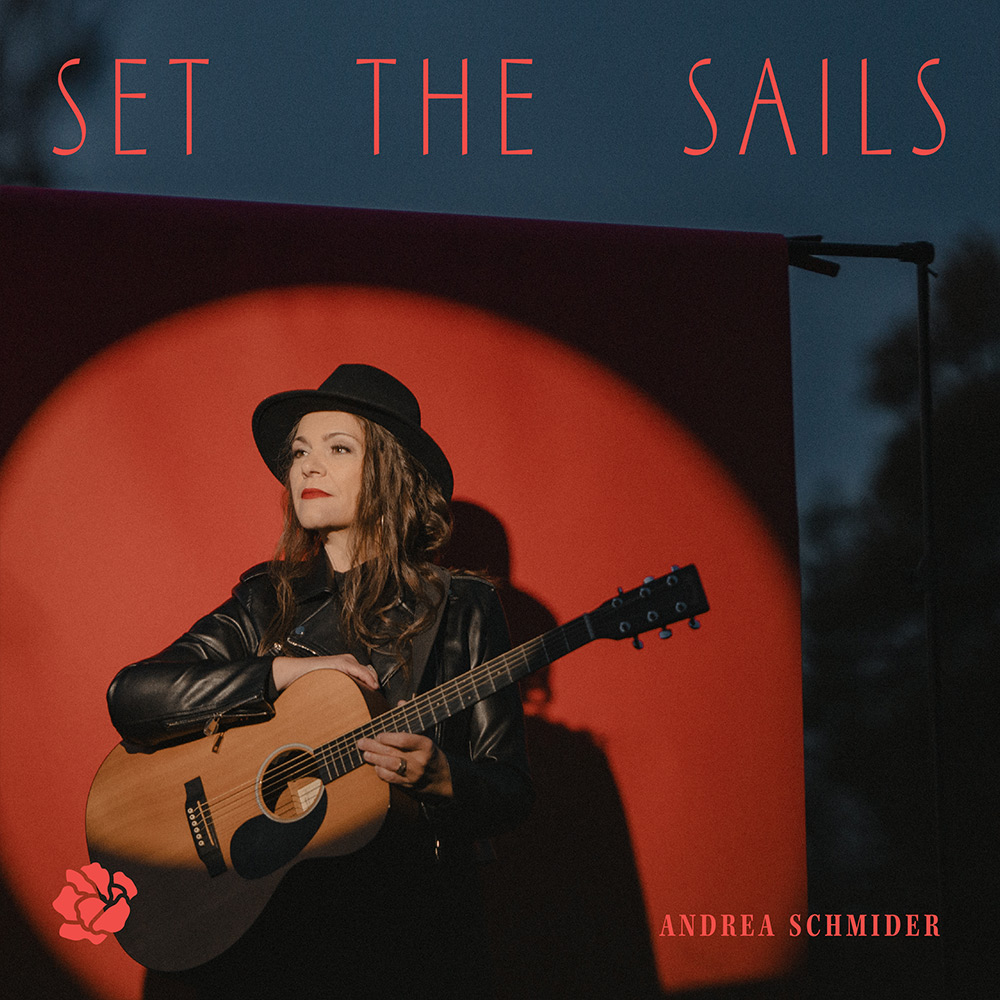 Andrea Schmider mit schwarzem Hut und Lederjacke, die eine Akustikgitarre vor rotem Hintergrund hält.