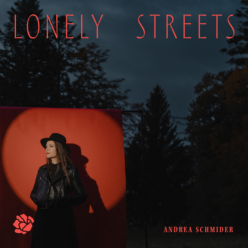 Frau in schwarzem Lederjacke und Hut vor rotem Hintergrund mit runden Licht, umgeben von dunklen Bäumen in der Dämmerung.