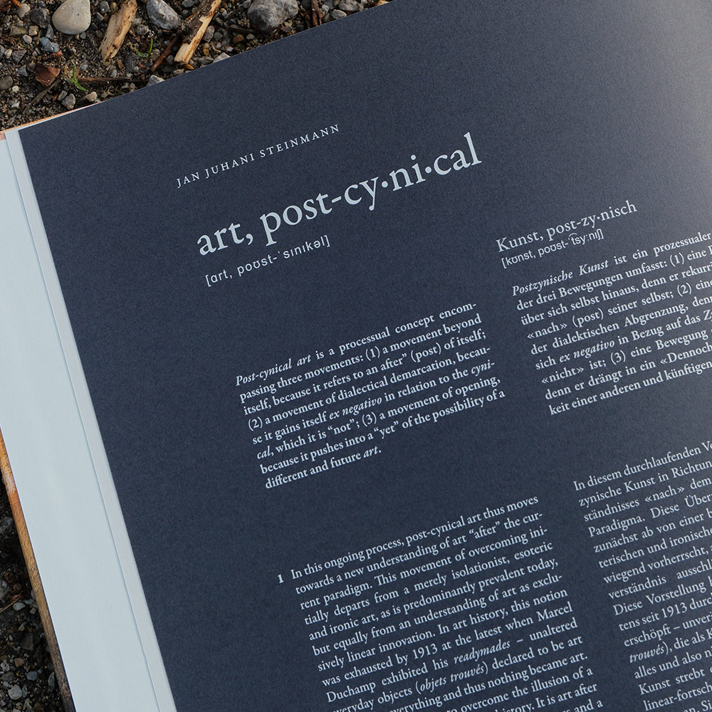 Foto eines aufgeschlagenen Buchs mit Architekturtext und Bild, auf einem erdigen, mit Moos bedeckten Boden liegend.