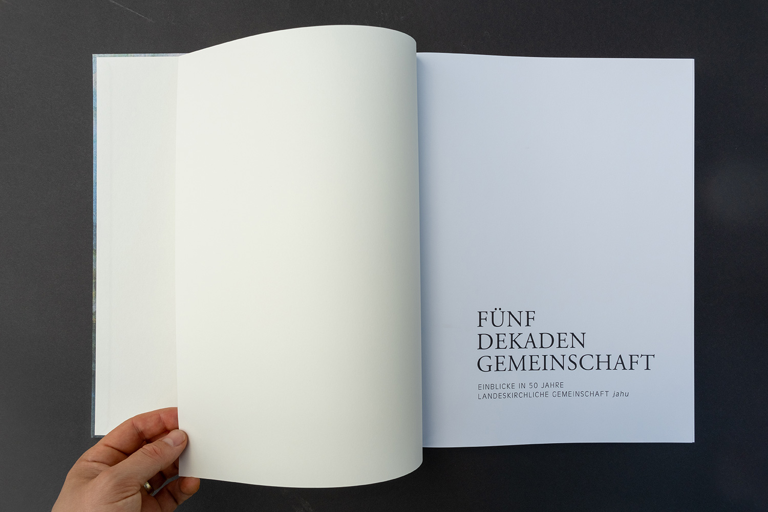 Hand blättert in Buch mit Titel 'Fünf Dekaden Gemeinschaft' und Untertitel auf weisser Seite.