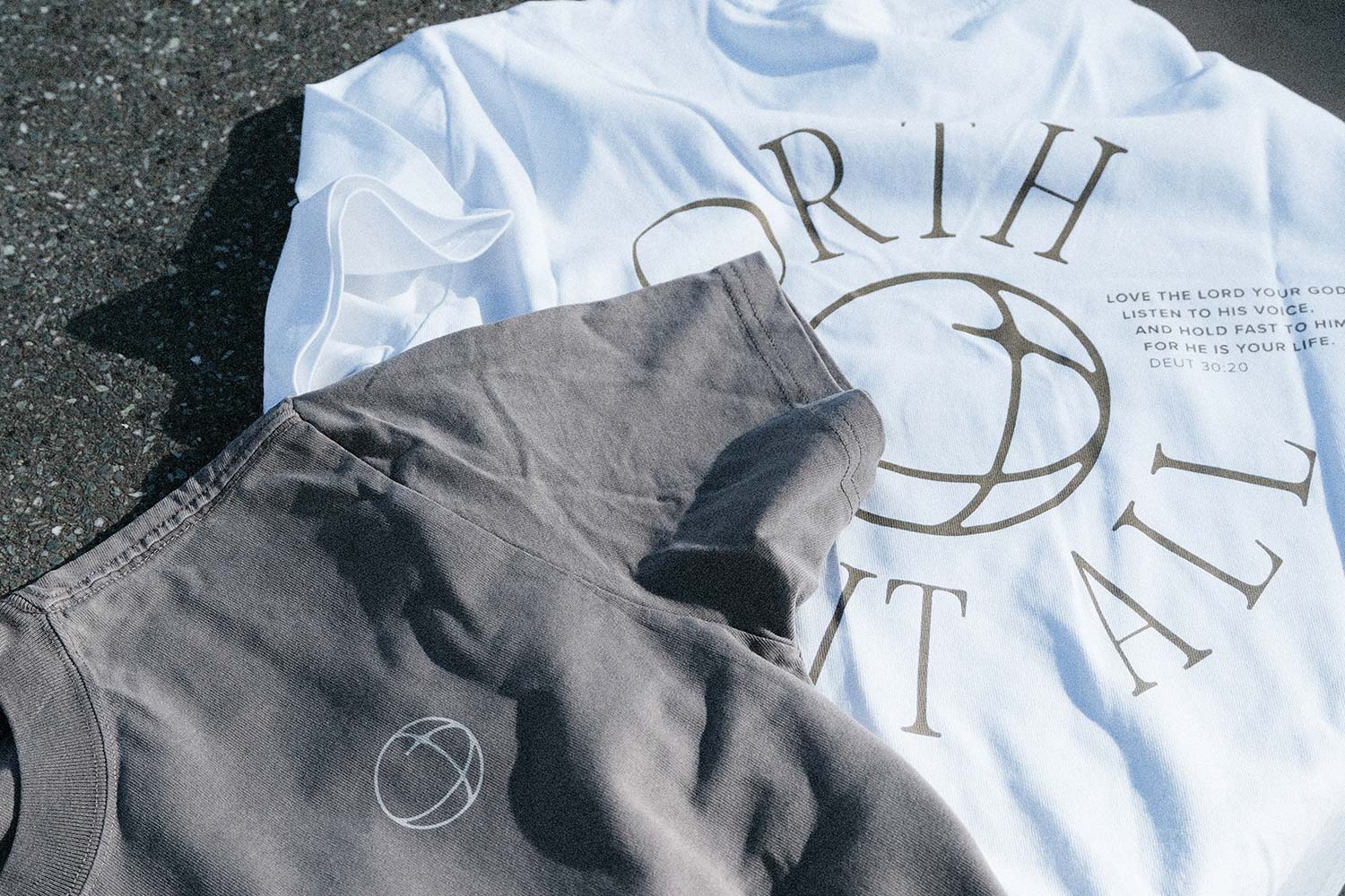 Weisses T-Shirt mit Schrift und graues T-Shirt mit kleinem Kreislogo nebeneinander auf Asphalt.