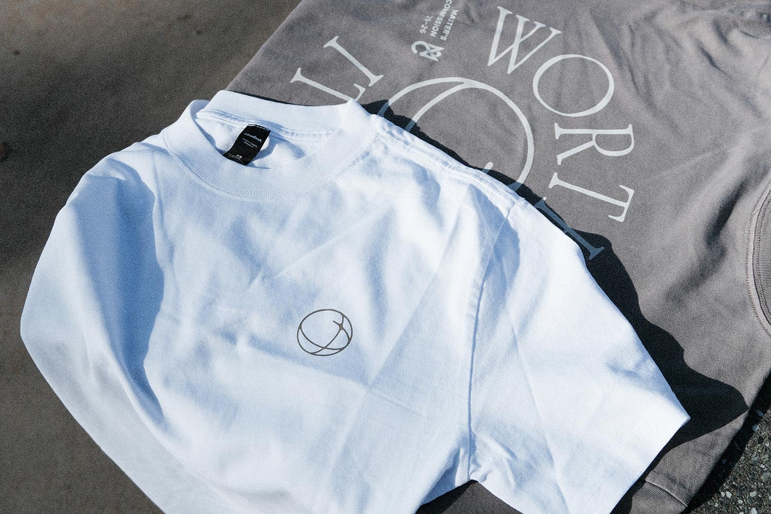Weißes T-Shirt mit kleinem rundem Logo neben einem grauen Shirt mit großem Textdruck.