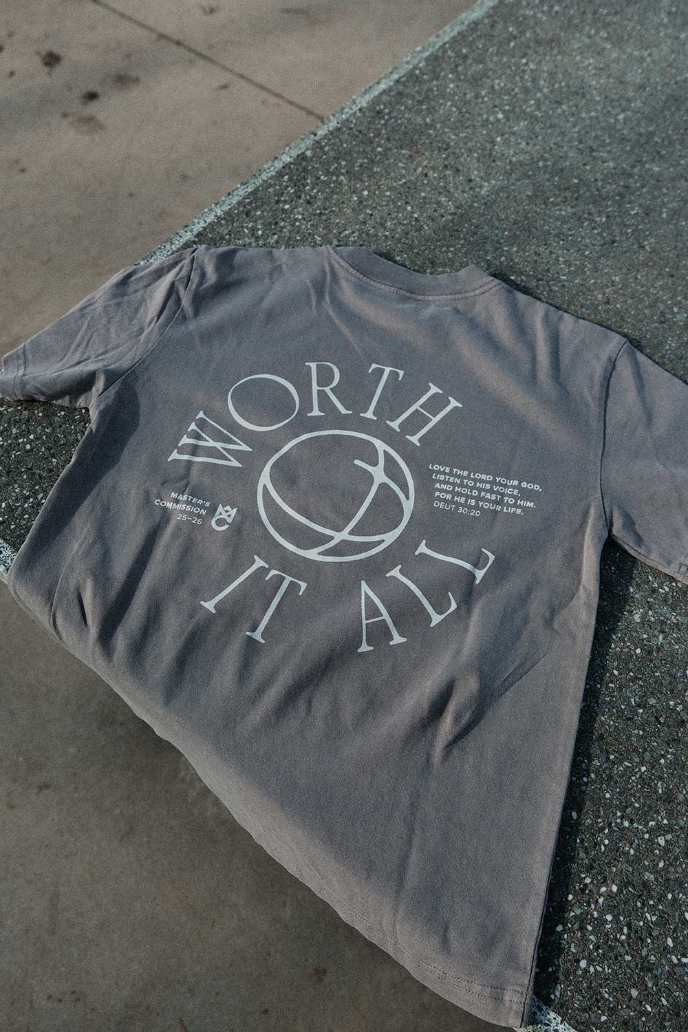 Grau T-Shirt mit weißem Schriftzug und einem Globus-Design, liegend auf einer steinernen Oberfläche.