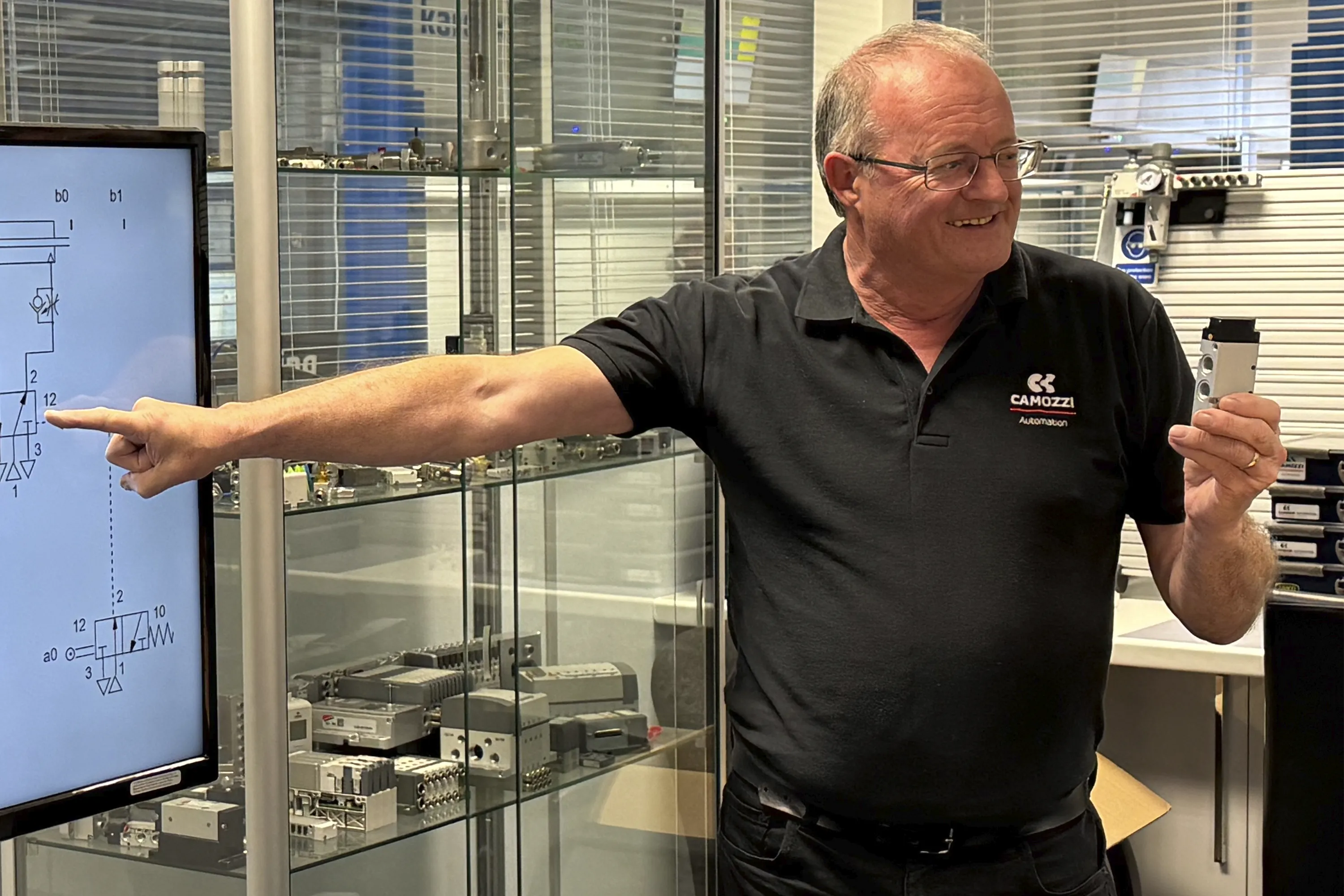 Steve Partington, bringing over 45 years of practical pneumatics experience into the classroom, smiles as he holds a physical valve while pointing to its corresponding ISO 1219 symbol on a digital circuit diagram. He is demonstrating how to connect mechanical theory to real-world applications inside the dedicated training facility at the Camozzi UK Automation Centre.