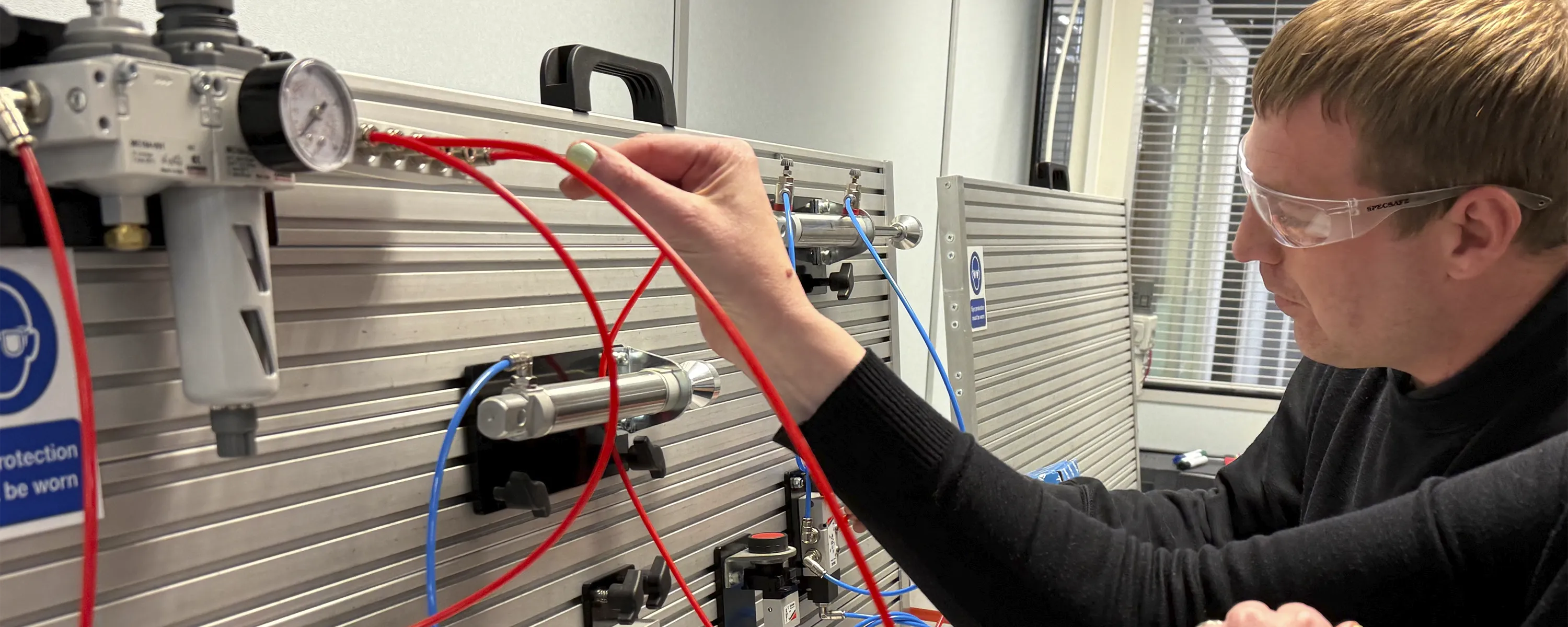 2 delegates wearing safety glasses connects red and blue tubing to a pneumatic training rig during a practical circuit building exercise at Camozzi Automation UK.