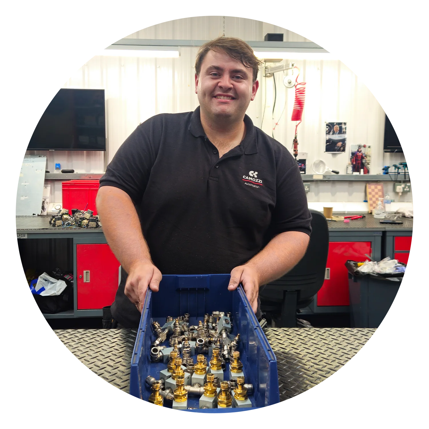 Jack McIntosh, Assembly Operative at Camozzi Automation UK, smiling while holding a blue parts bin filled with brass and metal pneumatic fittings at his workshop bench.