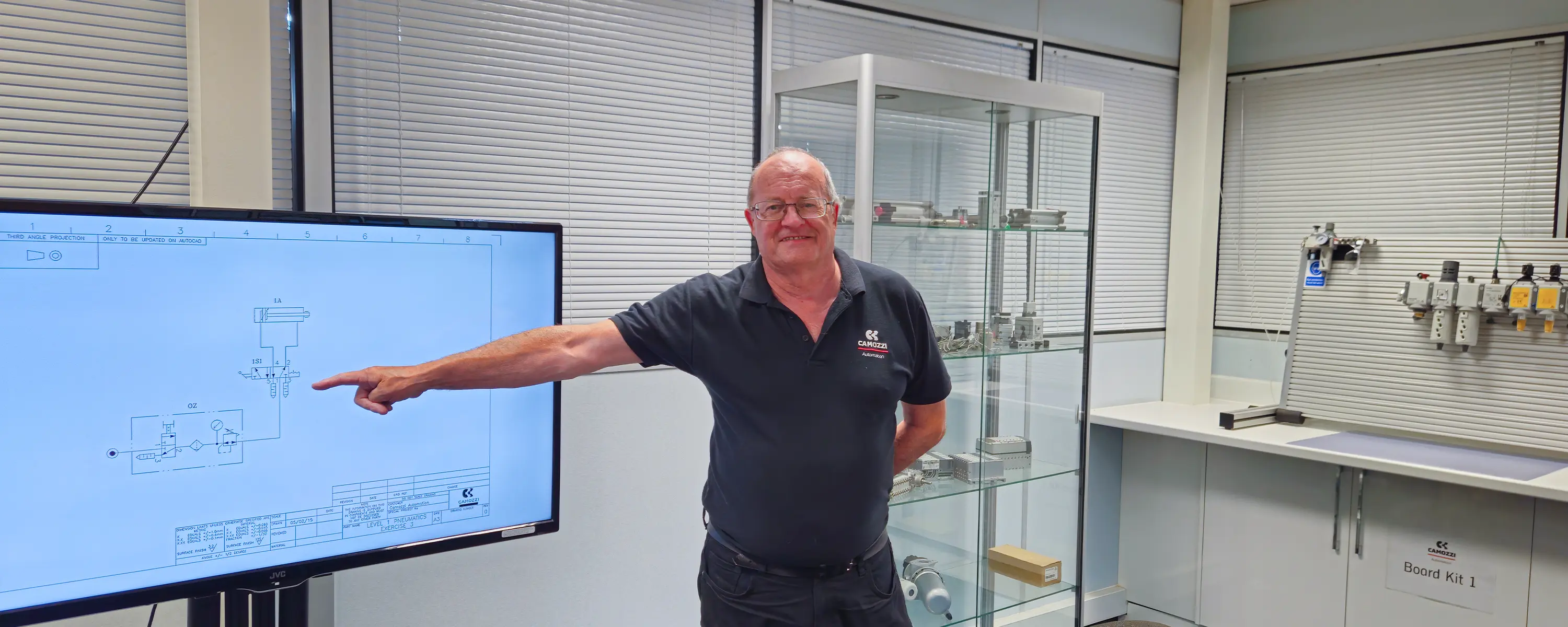 Steve Partington pointing to a pneumatic circuit drawing on a TV screen during a Camozzi Automation UK training session. A practical training board featuring an MX SAFEMAX unit and an FSX flow meter is visible in the background.