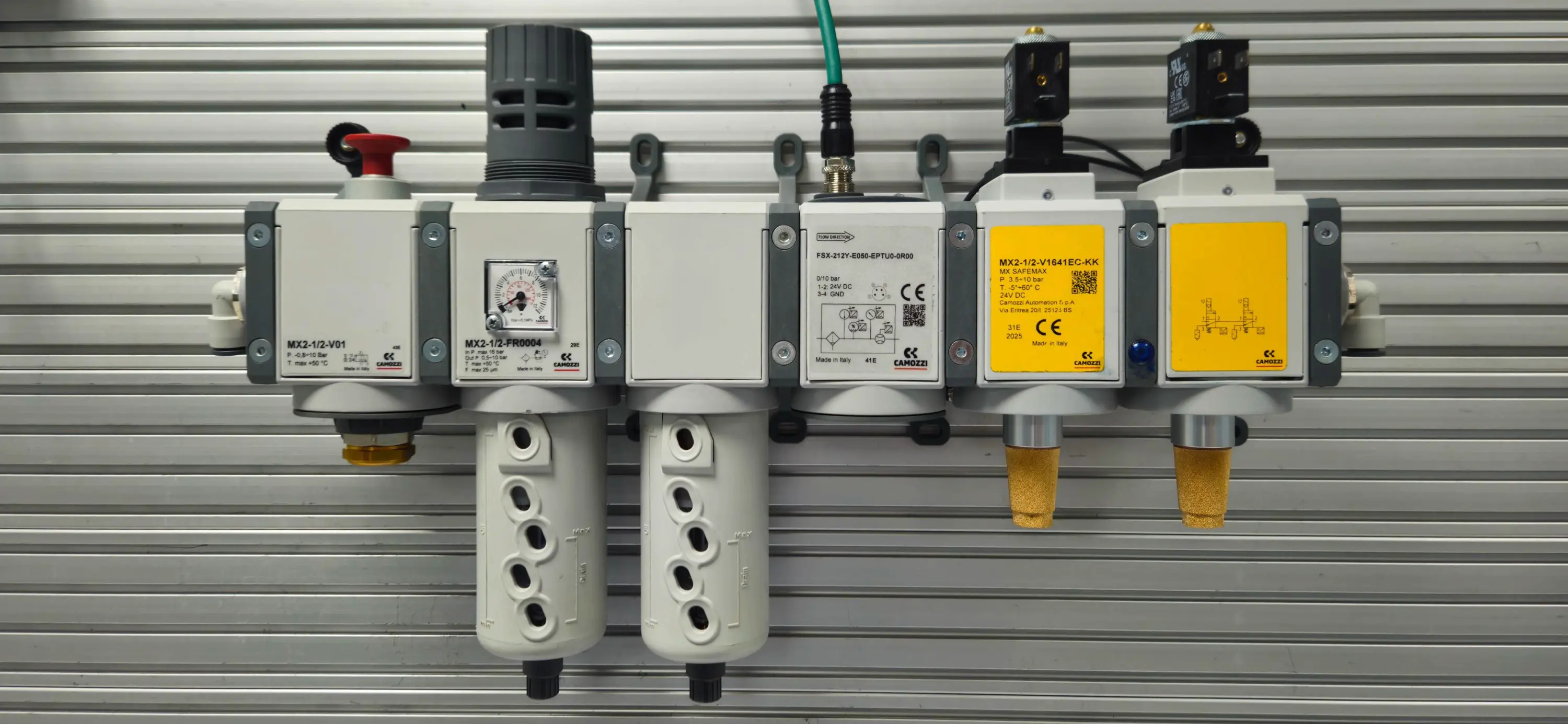 A hand holding the red Camozzi Pneu-Saver unit in front of a practical training board. The board displays a modular air preparation assembly, including an MX SAFEMAX safety unit and an FSX flow meter, used for the Camozzi UK Safety in Pneumatics and Energy Saving training course.