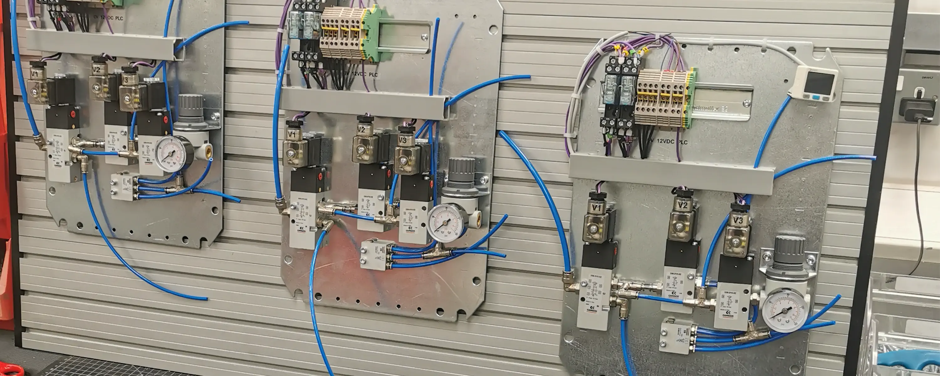 In progress assembly of custom electro-pneumatic control panels at the Camozzi UK Automation Centre, featuring metal backplates populated with solenoid valves, pressure regulators, electrical terminal blocks, and blue pneumatic tubing.