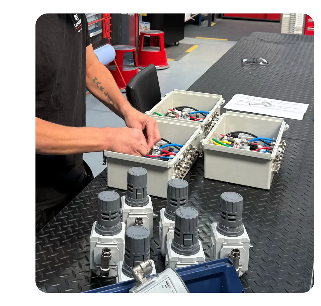 A Camozzi UK engineer assembling a batch of identical compact pneumatic control panels in grey enclosures on a workshop bench, with documentation and air regulators nearby.