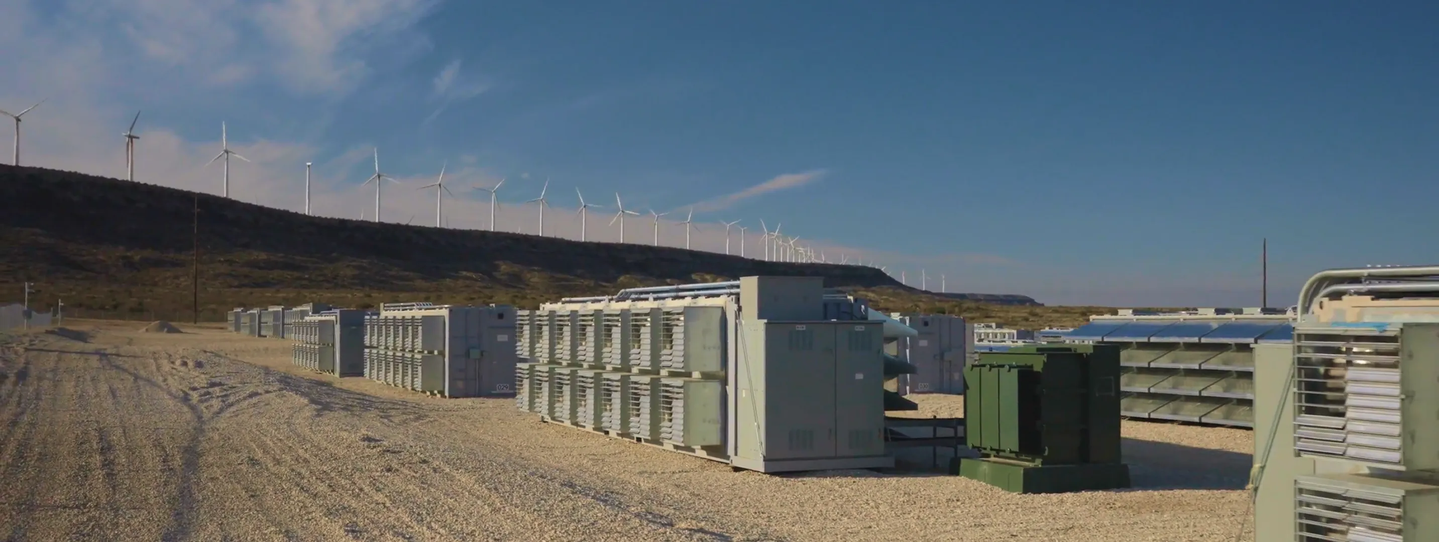 Rows of large industrial battery storage units on gravel with wind turbines on a hill in the background.
