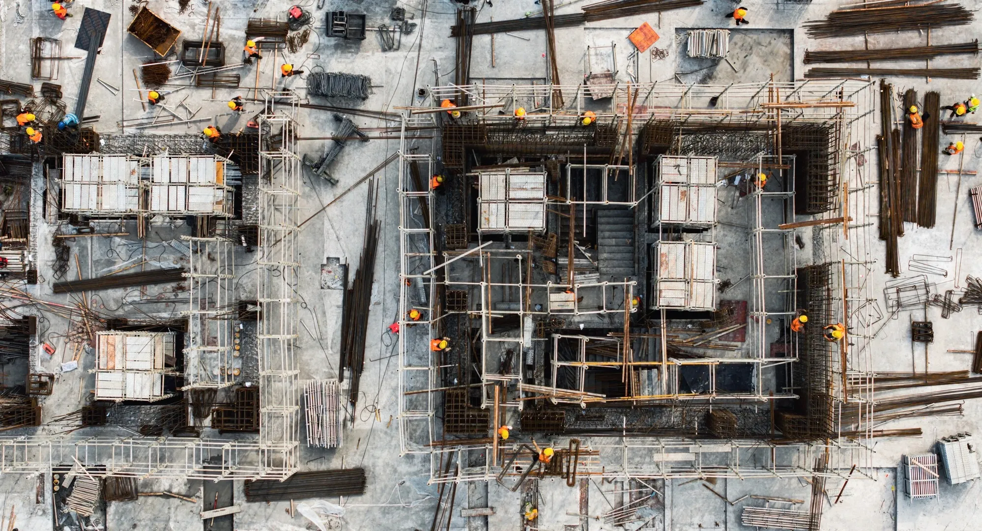 Overhead view of workers in yellow helmets at a large concrete construction site with scaffolding and steel rods.