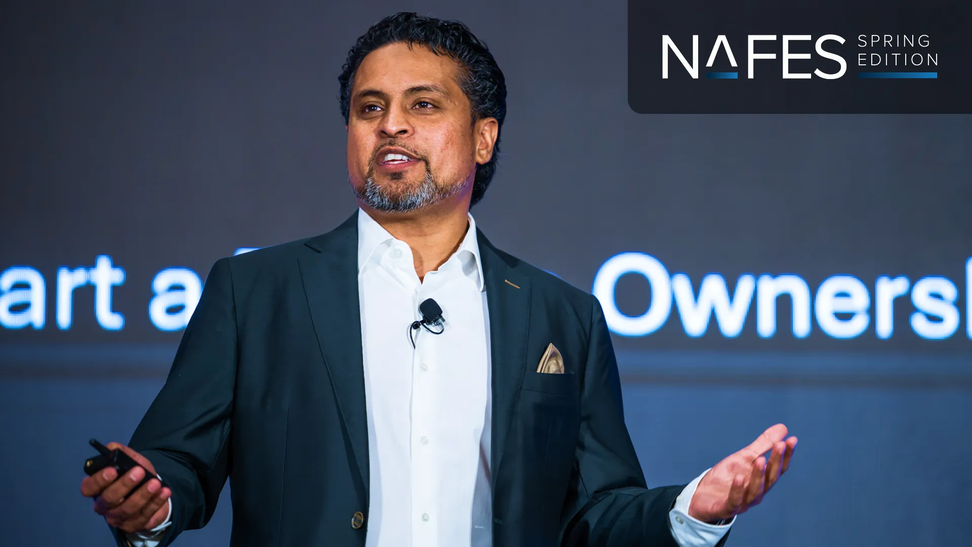 Man in suit speaking on stage with a microphone, gesturing with his hands, NAFES Spring Edition logo above.