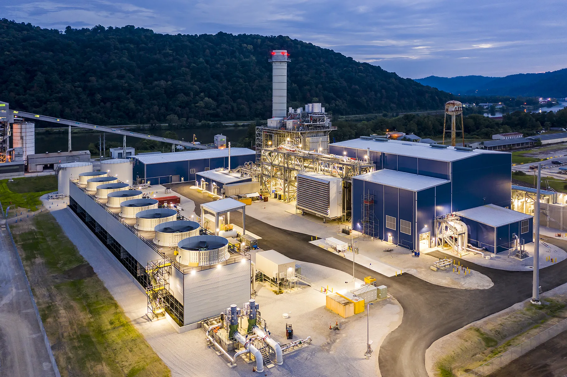 A large power plant facility with cooling towers and chimneys lit at dusk near forested hills.