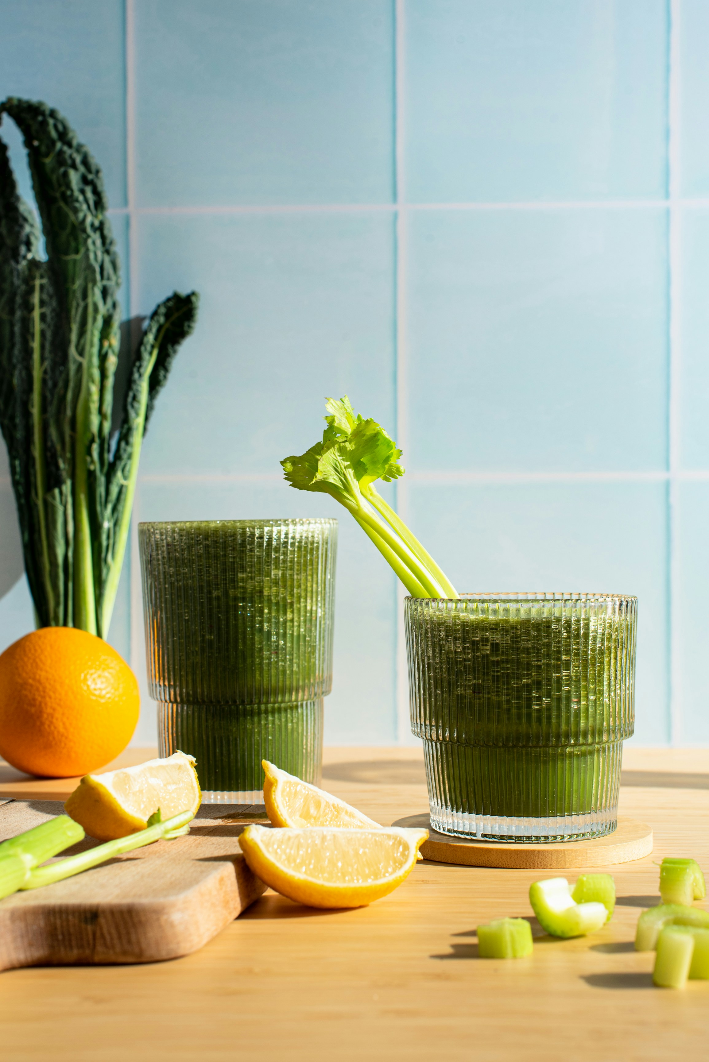 Two cups of green smoothies are on a table with slices of orange and celery spread around.