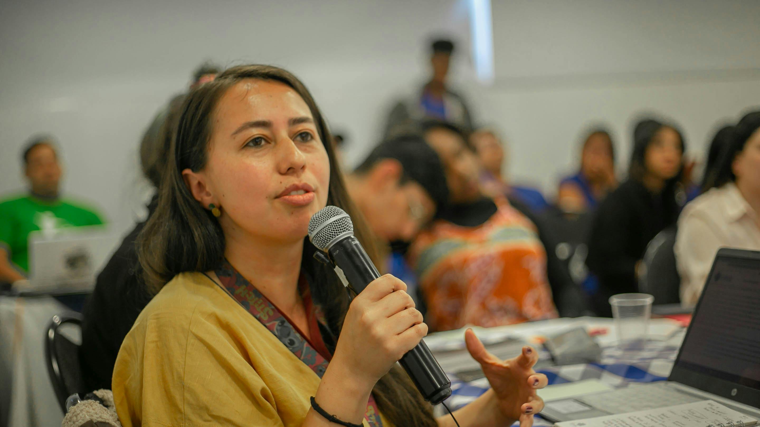 A woman in a room sat at a table holding a microphone with people around her.