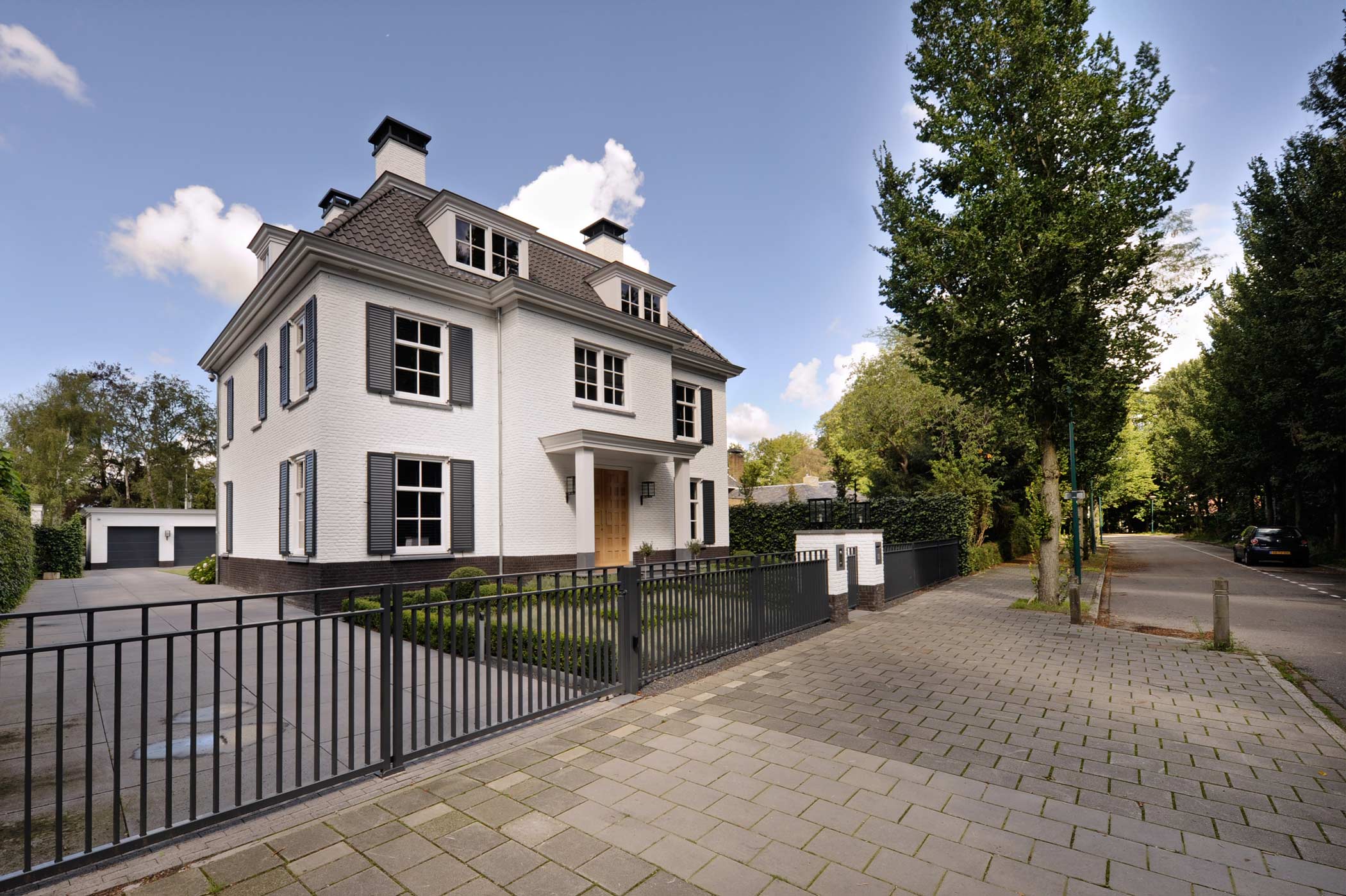 Groot wit huis met zwarte luiken, moderne zwarte hekwerk om tuin en oprit, op rustige straat.