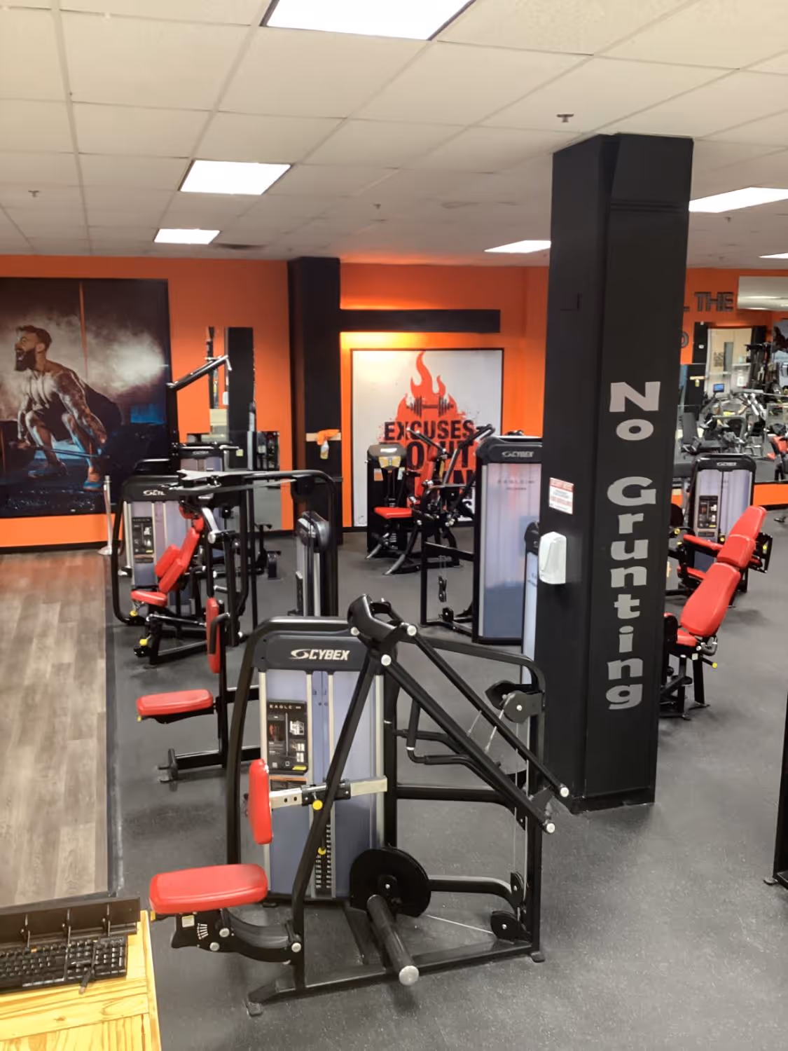 Interior of a gym with multiple Cybex weight machines with red seats, an orange wall, and a motivational sign that reads 'Excuses don't burn calories'.