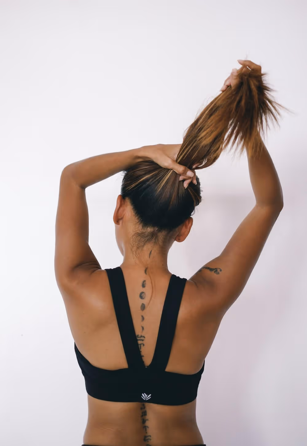Woman in black sports bra tying her long hair up, showing tattoos on her back and arm.