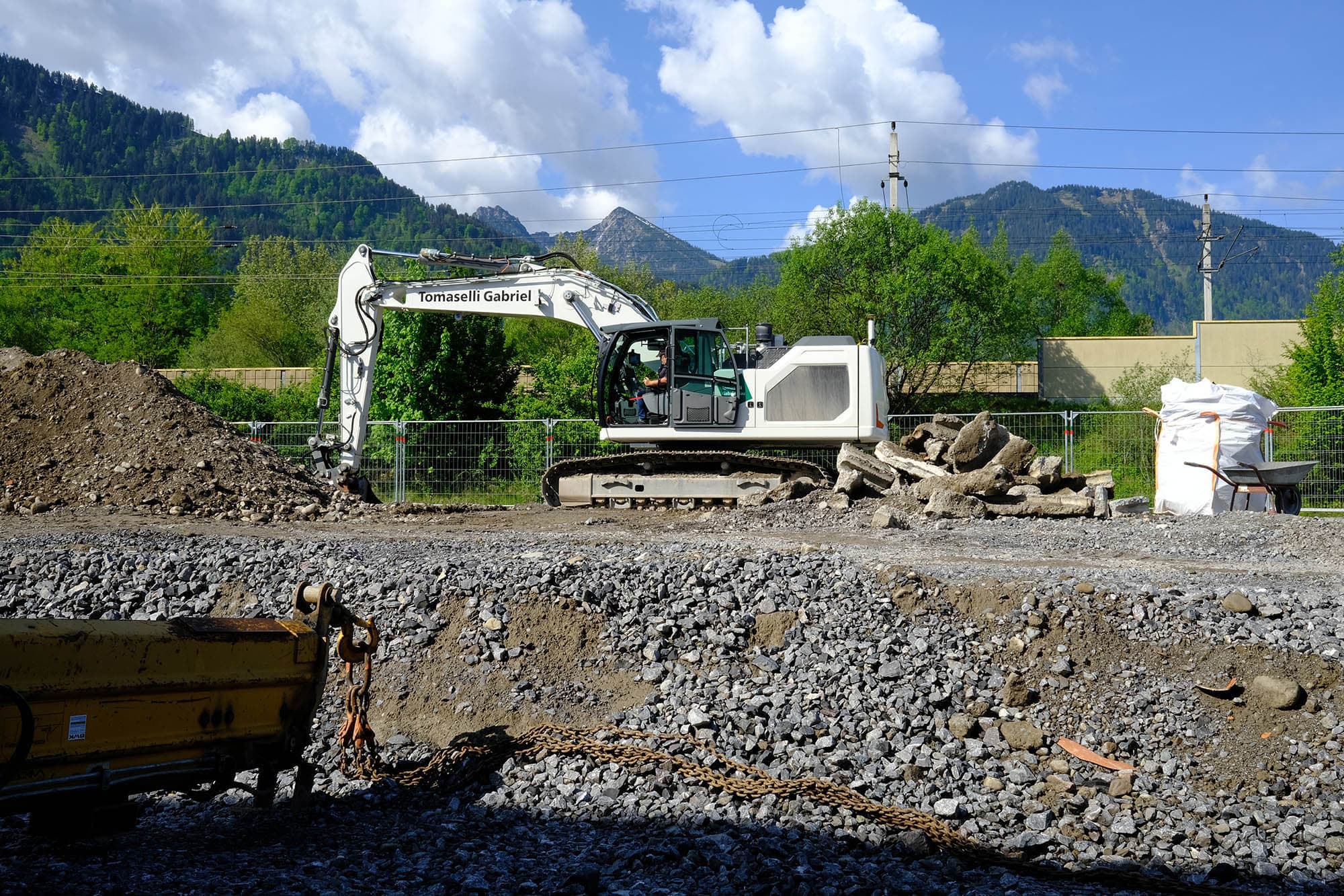 White excavator with company logo of Tomaselli Gabriel, in the foreground excavation pit, in the background mountains.