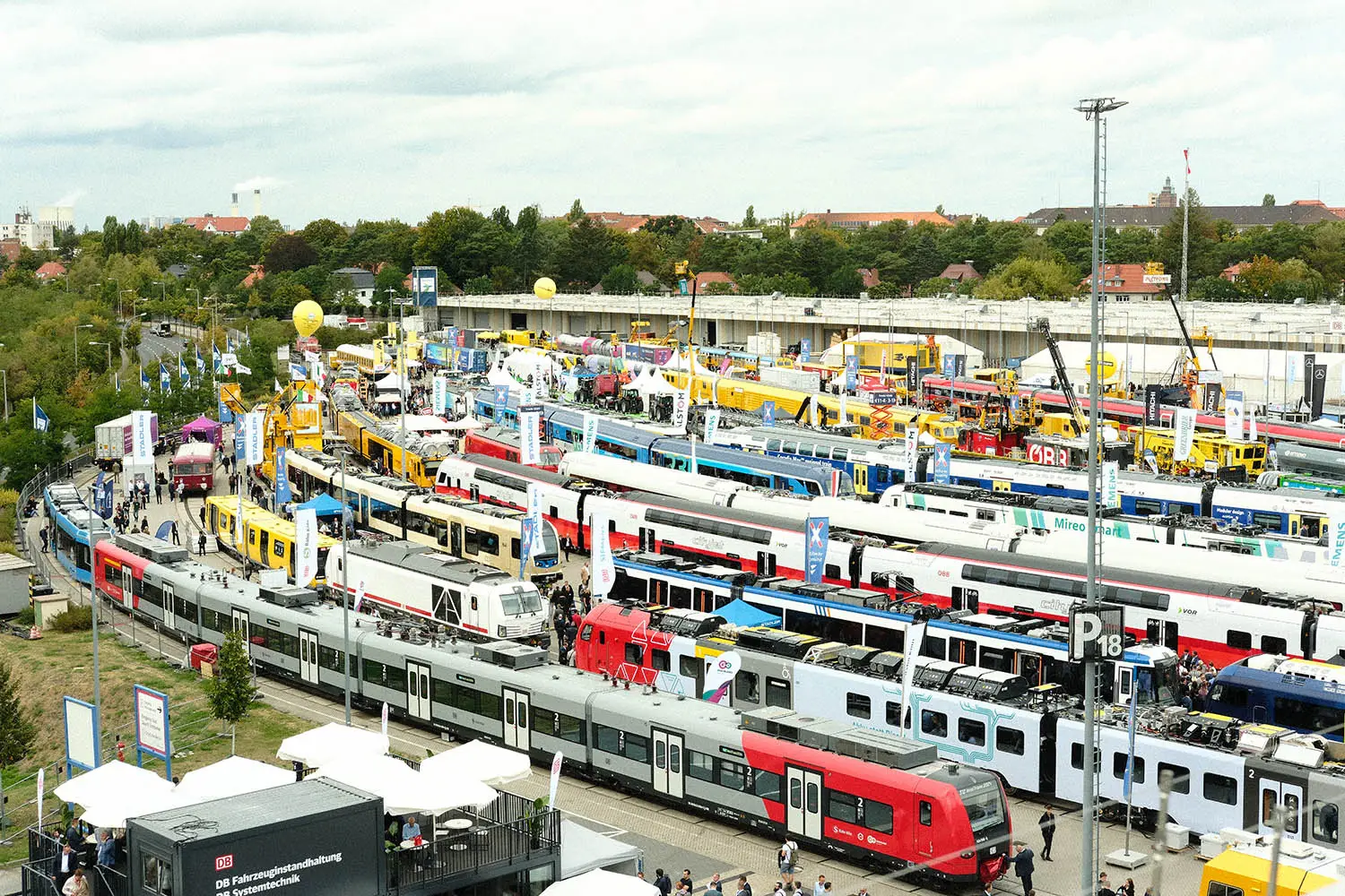 Outdoor railway trade show with multiple modern trains, booths, cranes, and crowds in a rail yard.