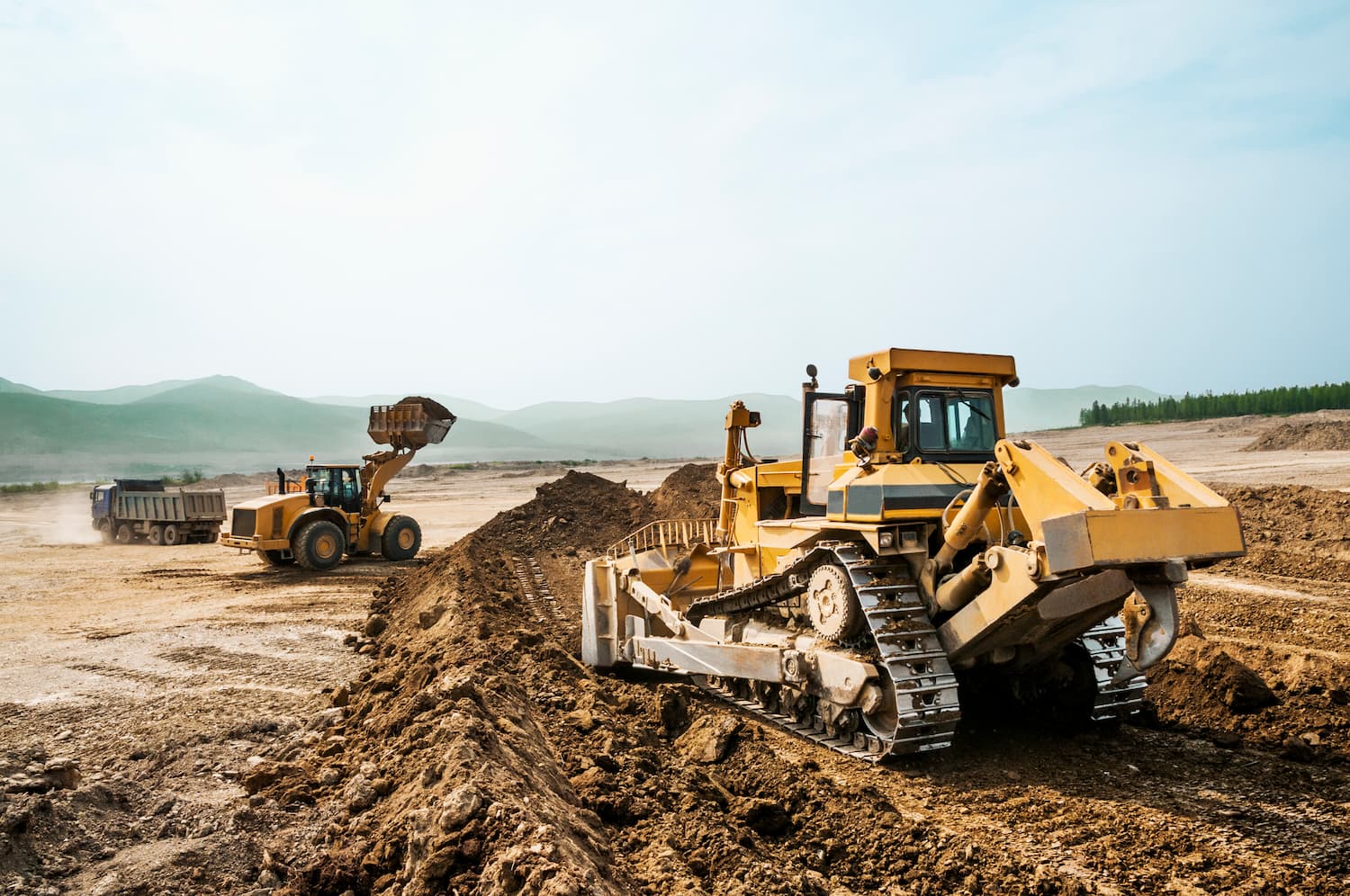 A large yellow bulldozer is pushing a mound of dirt across a wide, dusty construction site. Behind it, a wheel loader.