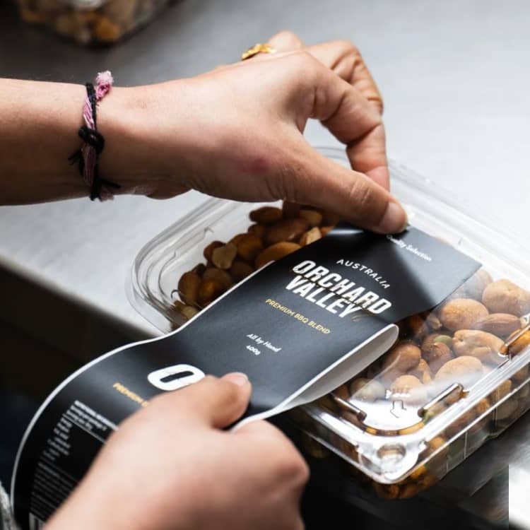 Orchard Valley sticker being applied to clear plastic container filled with farm fresh nuts