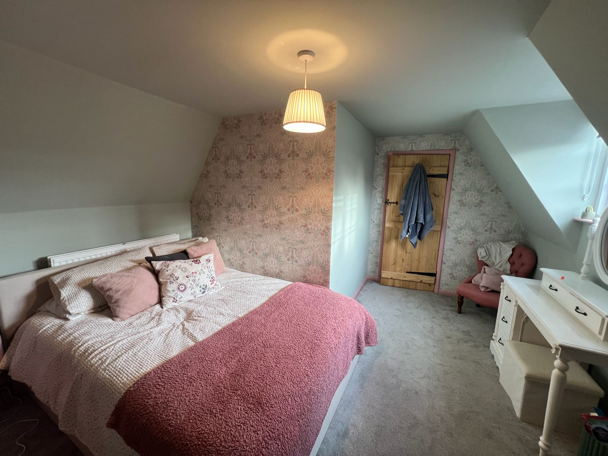 Cozy bedroom with floral wallpaper, a bed with pink and white bedding, a wooden door, pink cushioned chair, and white vanity table under a slanted ceiling.