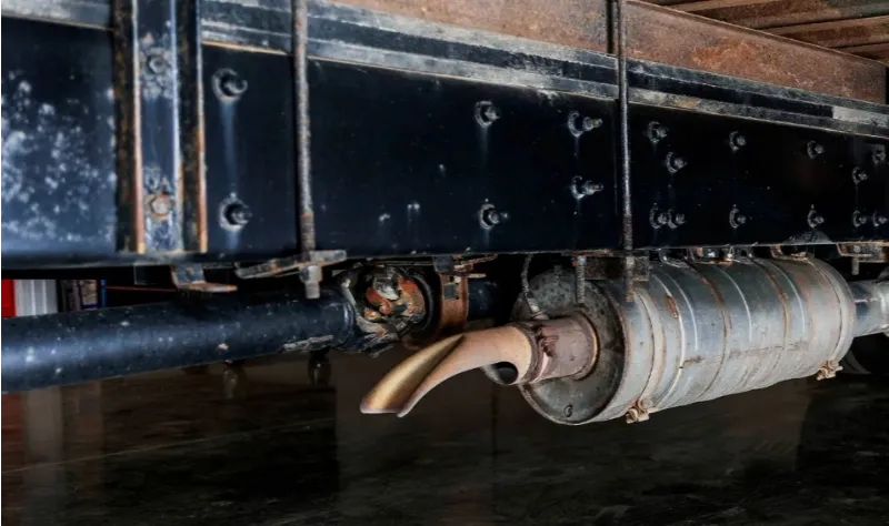 Exhaust system on a heavy-duty truck showing a rusted muffler and tailpipe beneath the chassis.