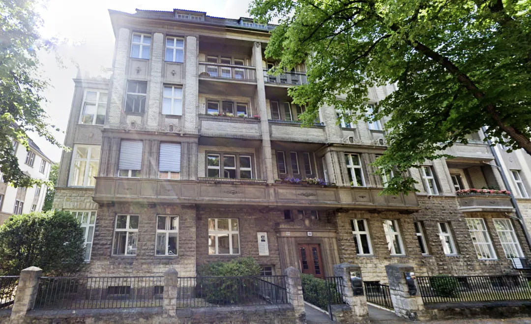 Außenansicht eines Bürogebäudes in Berlin bei Tageslicht. Das einstöckige Gebäude hat eine Glasfassade mit großen Fenstern, durch die man teilweise ins Innere sehen kann. Vor dem Bürogebäude stehen mehrere Autos geparkt. Im Hintergrund sind Bäume mit frischem grünen Laub zu sehen, die auf den Frühling hinweisen. Das Wetter ist klar und sonnig.