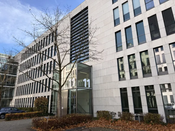 Ein dreistöckiges weißes Gebäude mit zahlreichen Fenstern, von denen einige geöffnet und andere geschlossen sind. Vor dem Gebäude befindet sich ein asphaltierter Parkplatz mit wenigen geparkten Autos. Der Himmel ist blau mit einigen weißen Wolken. In der linken unteren Ecke ist ein Eingang mit einem Vordach zu sehen. Im Gebäude befindet sich das Büro von Zenveo