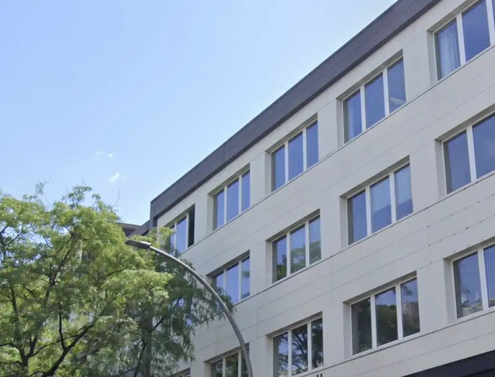 Außenansicht eines Bürogebäudes in Berlin bei Tageslicht. Das einstöckige Gebäude hat eine Glasfassade mit großen Fenstern, durch die man teilweise ins Innere sehen kann. Vor dem Bürogebäude stehen mehrere Autos geparkt. Im Hintergrund sind Bäume mit frischem grünen Laub zu sehen, die auf den Frühling hinweisen. Das Wetter ist klar und sonnig.