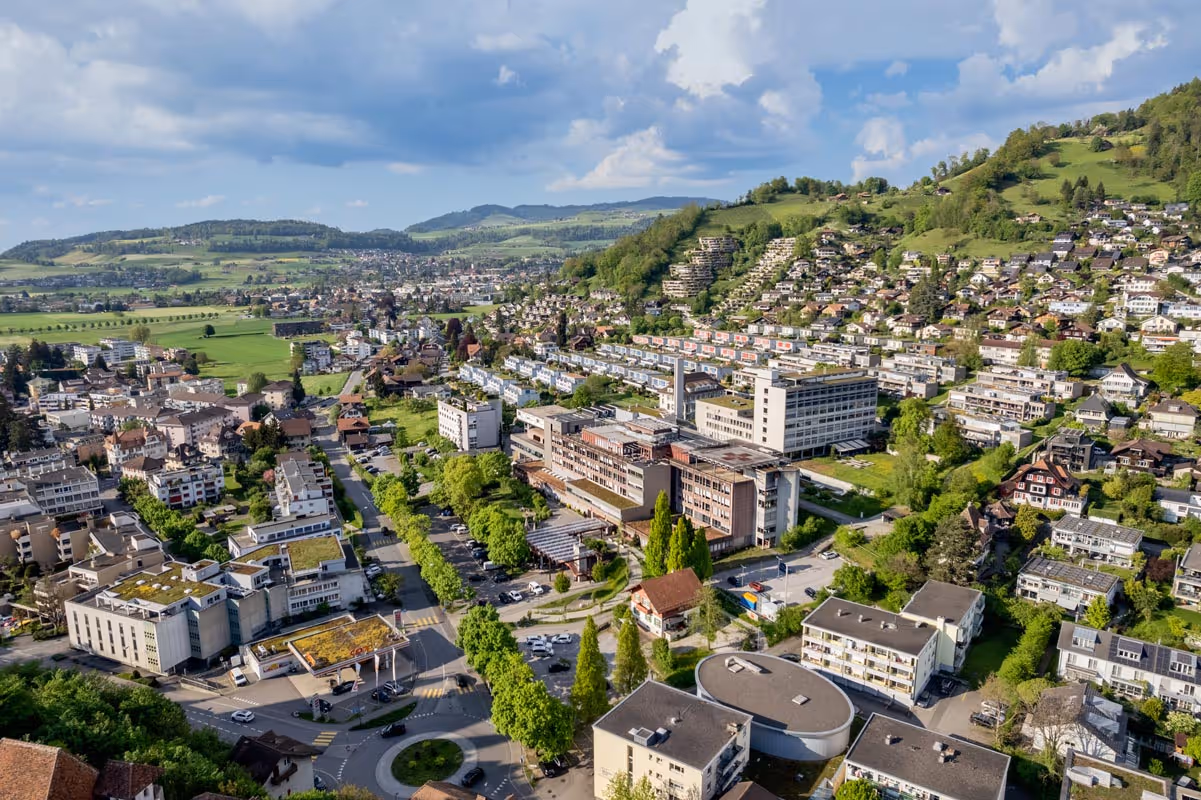 Luftaufnahme einer Stadt mit Wohngebieten, modernen Gebäuden und grünen Hügeln unter einem teils bewölkten Himmel.