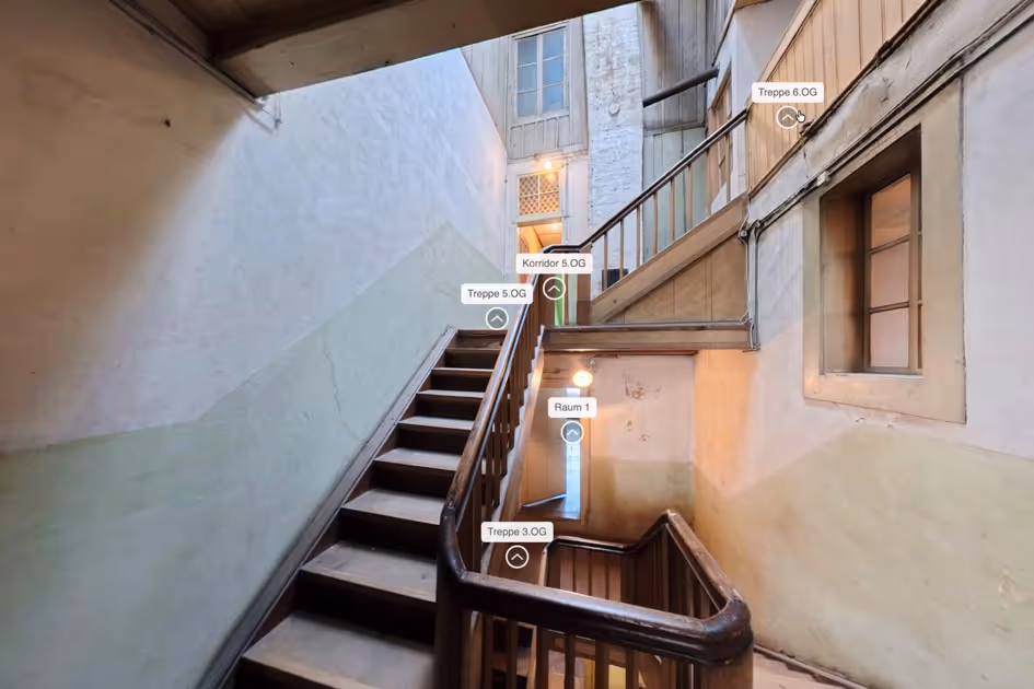 Alter hölzerner Treppenaufgang in einem Gebäude mit beigen Wänden und Beschriftungen zu verschiedenen Stockwerken und Räumen.