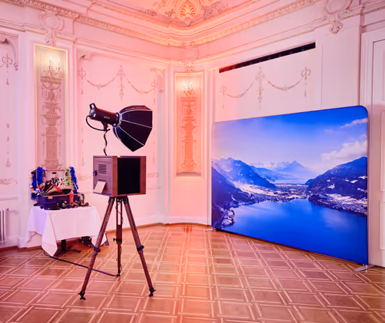 Klassisch eingerichteter Raum mit Fotobox auf Stativ, beleuchtet von Studiolicht, neben einem Tisch mit Requisiten und einem großen Panorama-Hintergrundbild der Schweizer Alpen.