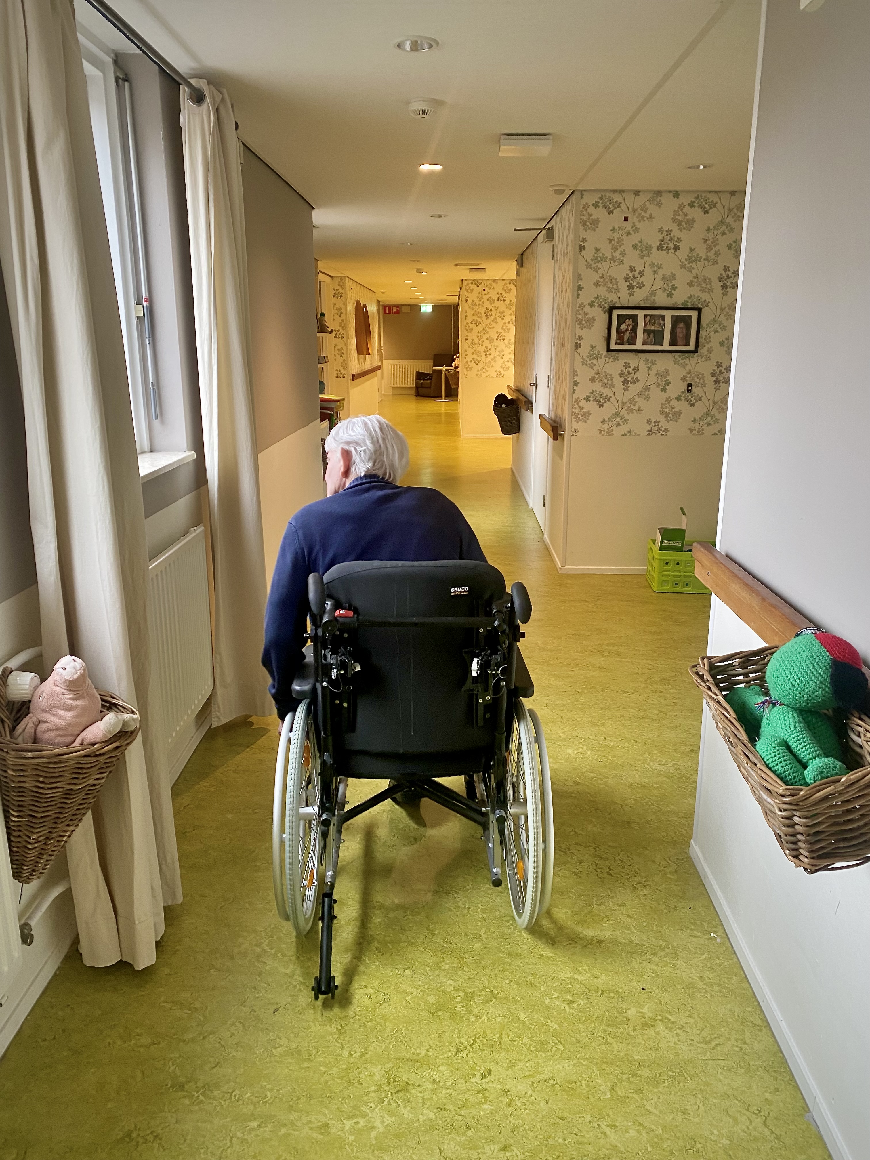 Een man in een rolstoel in de gang van een zorginstelling, van achteren gezien