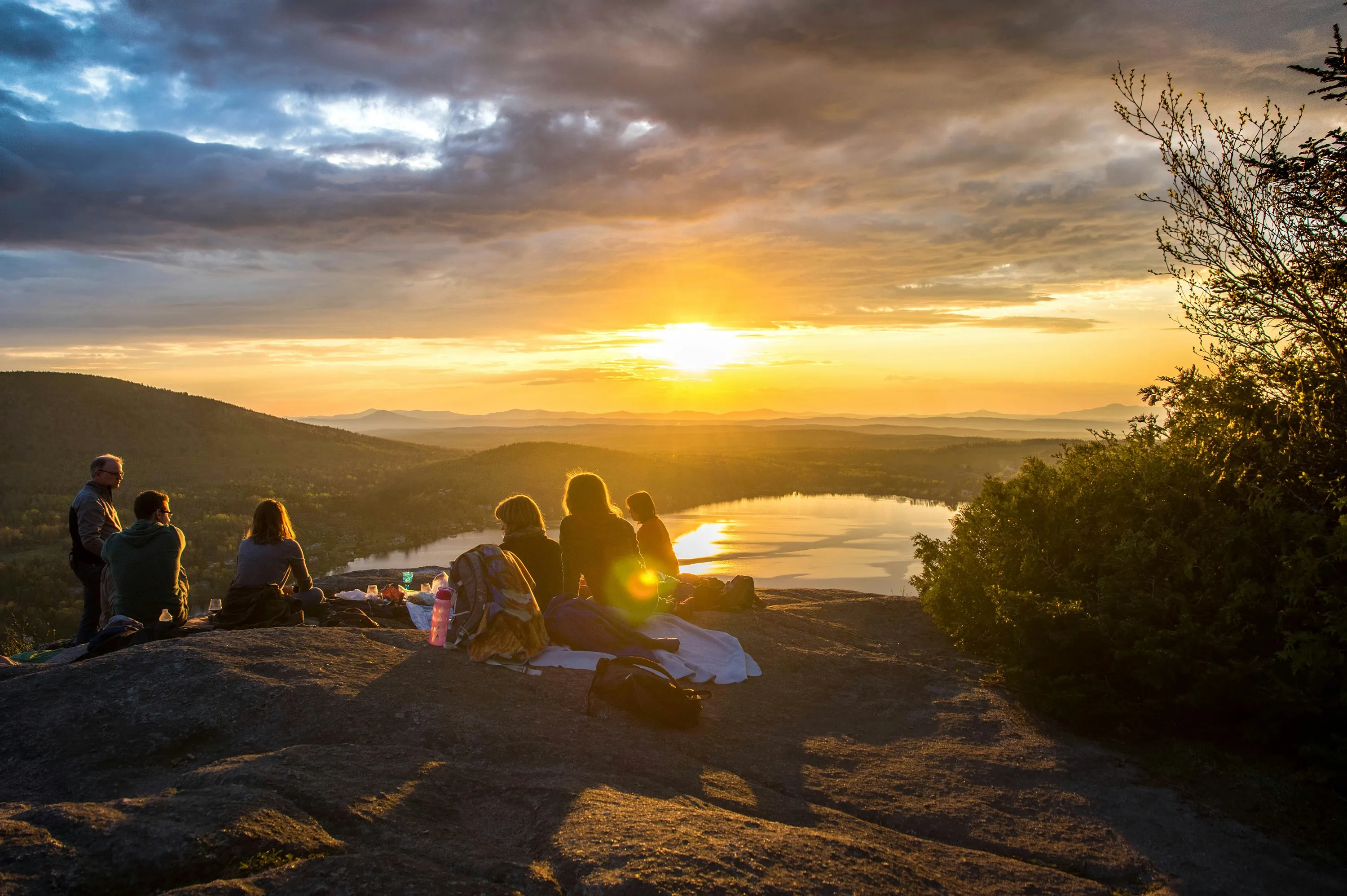 Group of people sitting on a rocky hilltop watching a sunset over a lake and distant mountains.