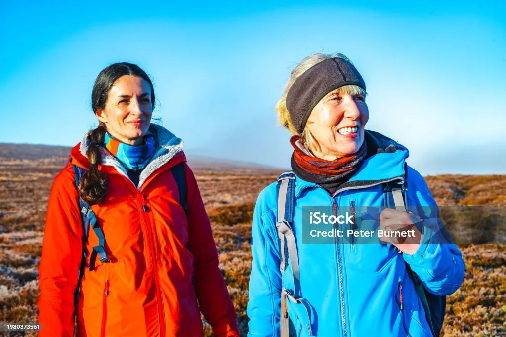 Two women hiking outdoors in cold weather, one wearing a red jacket and the other a blue jacket and headband, standing on a barren landscape.