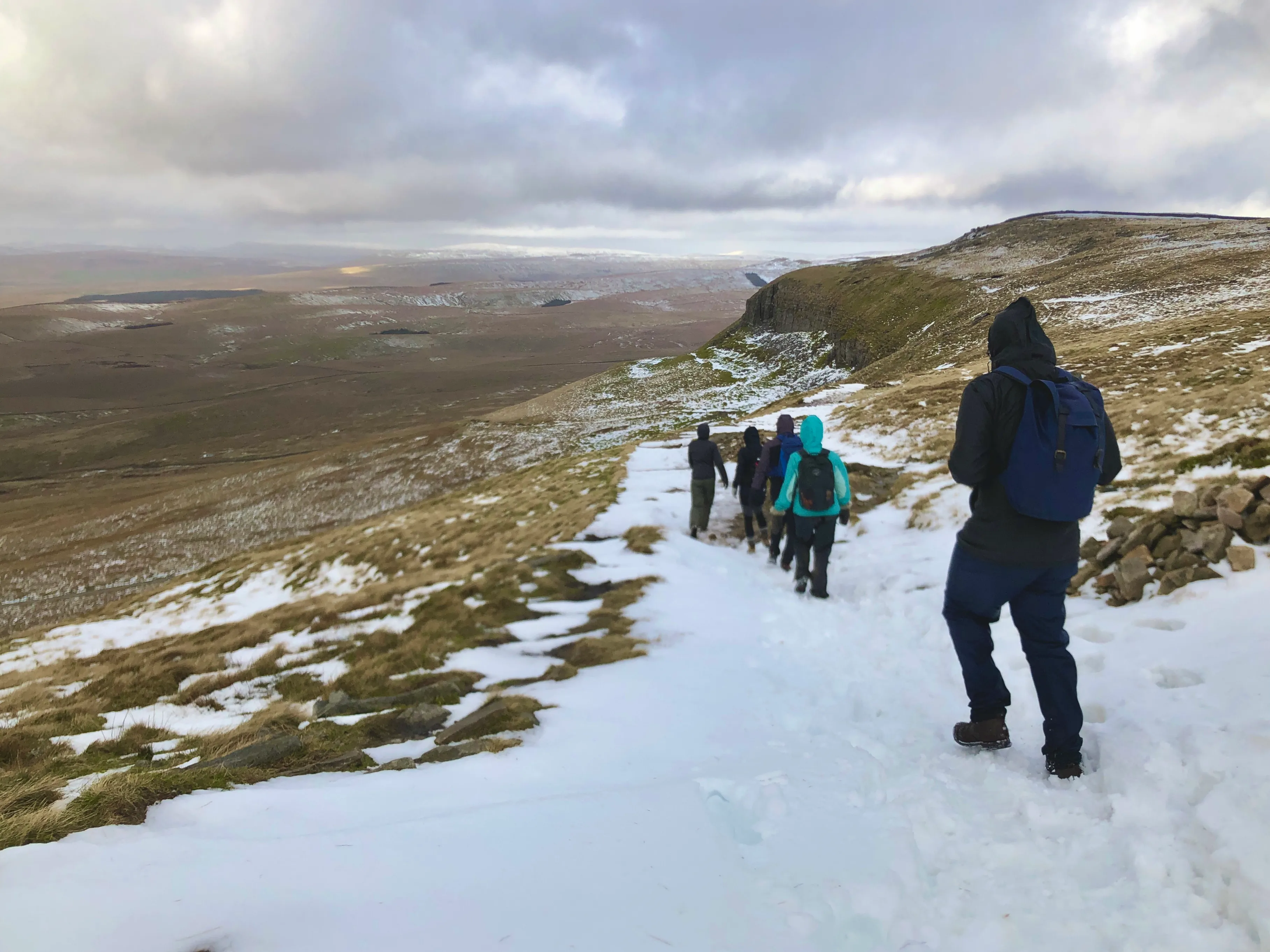 Group of hikers walking on a snowy path through a hilly landscape under a cloudy sky.