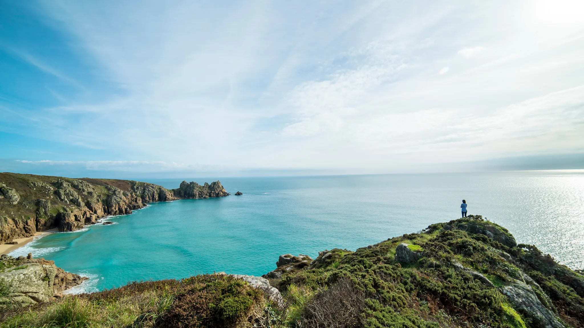 Person standing on a rocky, grassy cliff overlooking turquoise ocean water and distant rocky coastline under a partly cloudy sky.