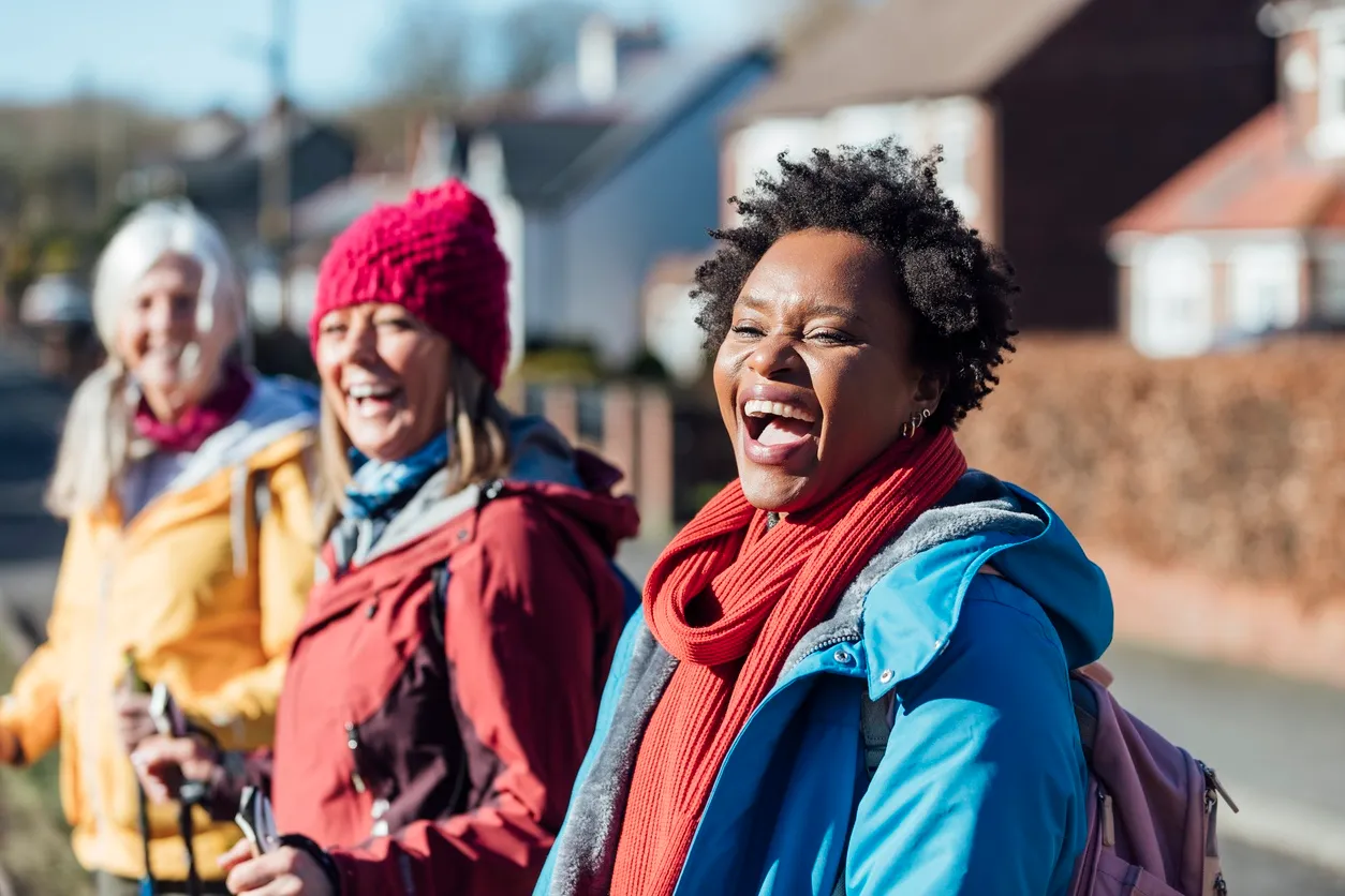 Three women dressed in winter clothing smiling and laughing outdoors on a sunny day.