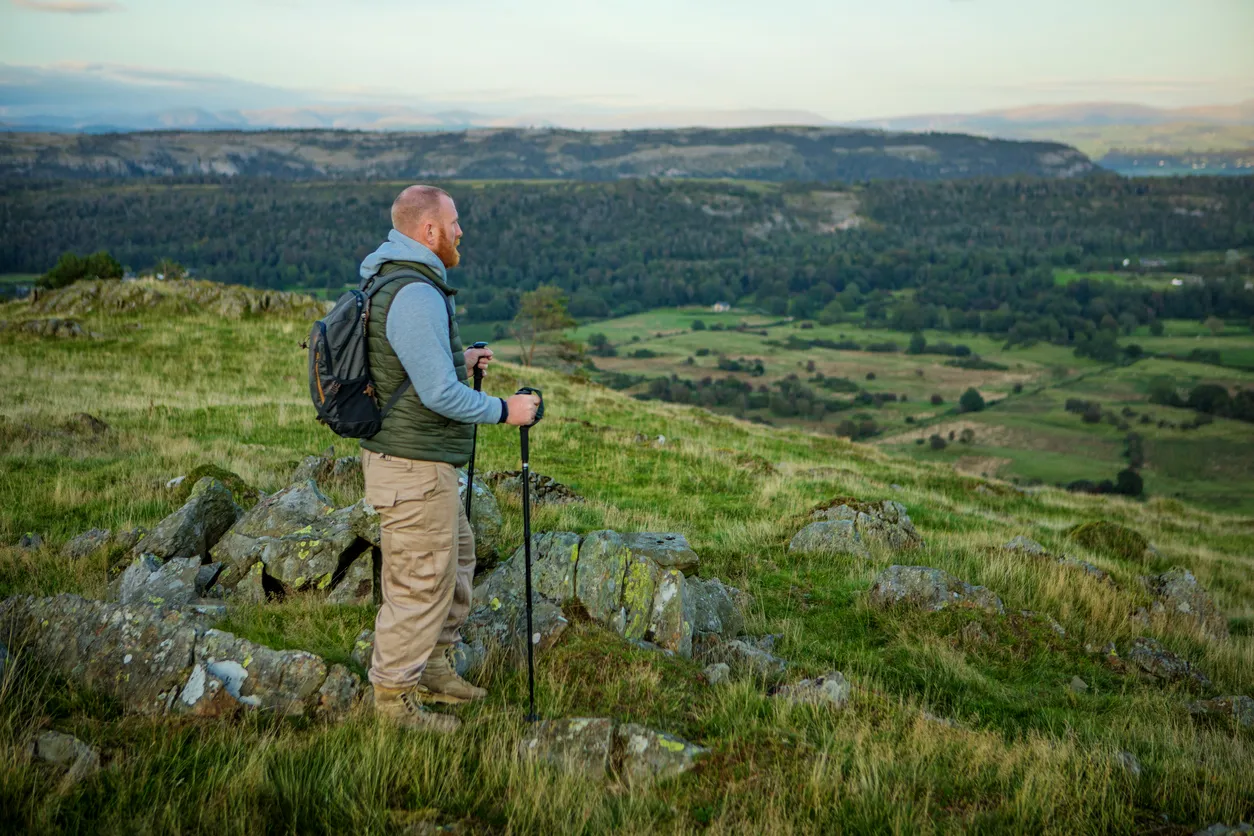 Hiker with backpack and trekking poles standing on rocky terrain overlooking a green valley and distant hills.