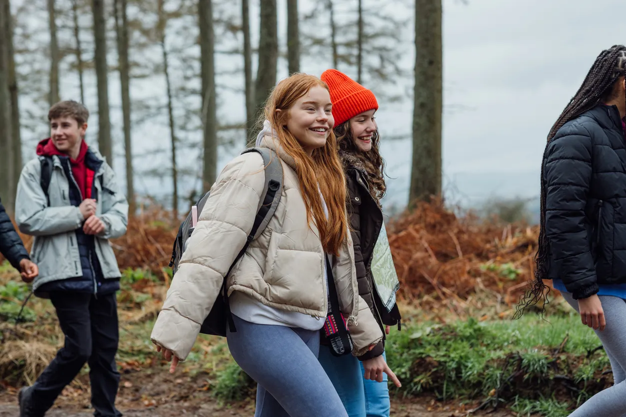 Group of young people walking and smiling outdoors in a forested area wearing winter jackets.