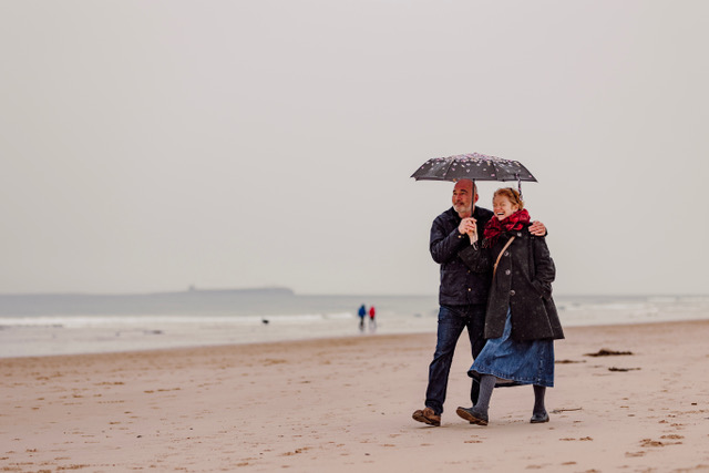 A couple laughing and walking along the beach enjoying the coastal margin, holding an umbrella, holding hands in the rain